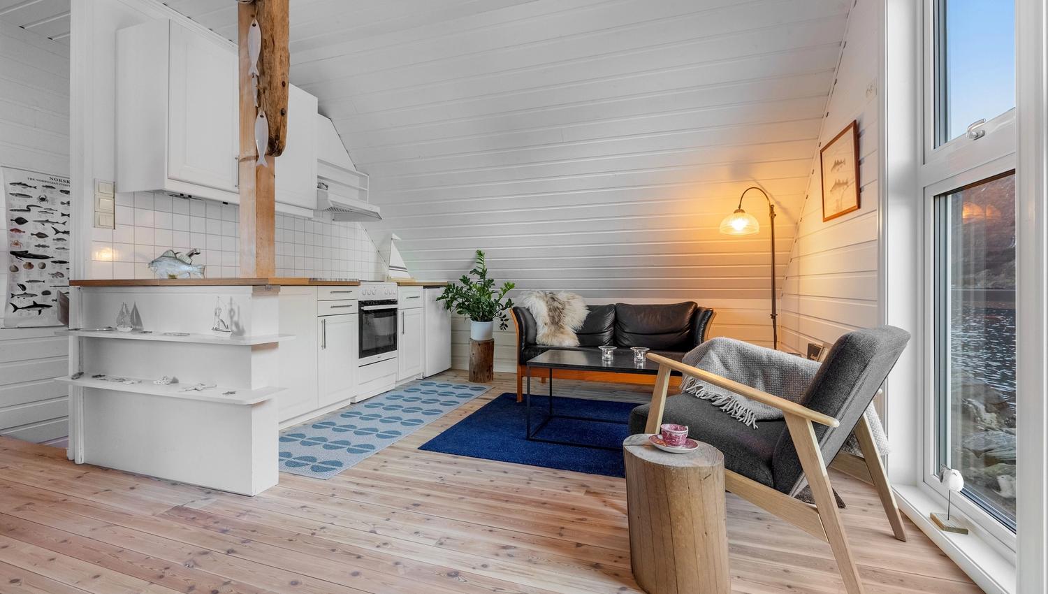 Bright wood-paneled cabin interior with white kitchen, sofa, armchair, coffee table, and large window by the sea at the Seahouse at Hornelen Panorama in Bremanger..