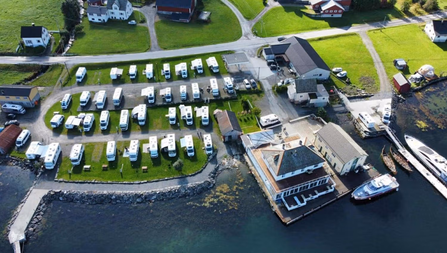 Aerial view over Davik Fjordparadis in Bremanger of a seaside RV campground with rows of white campers, nearby buildings, roads, and docked boats..
