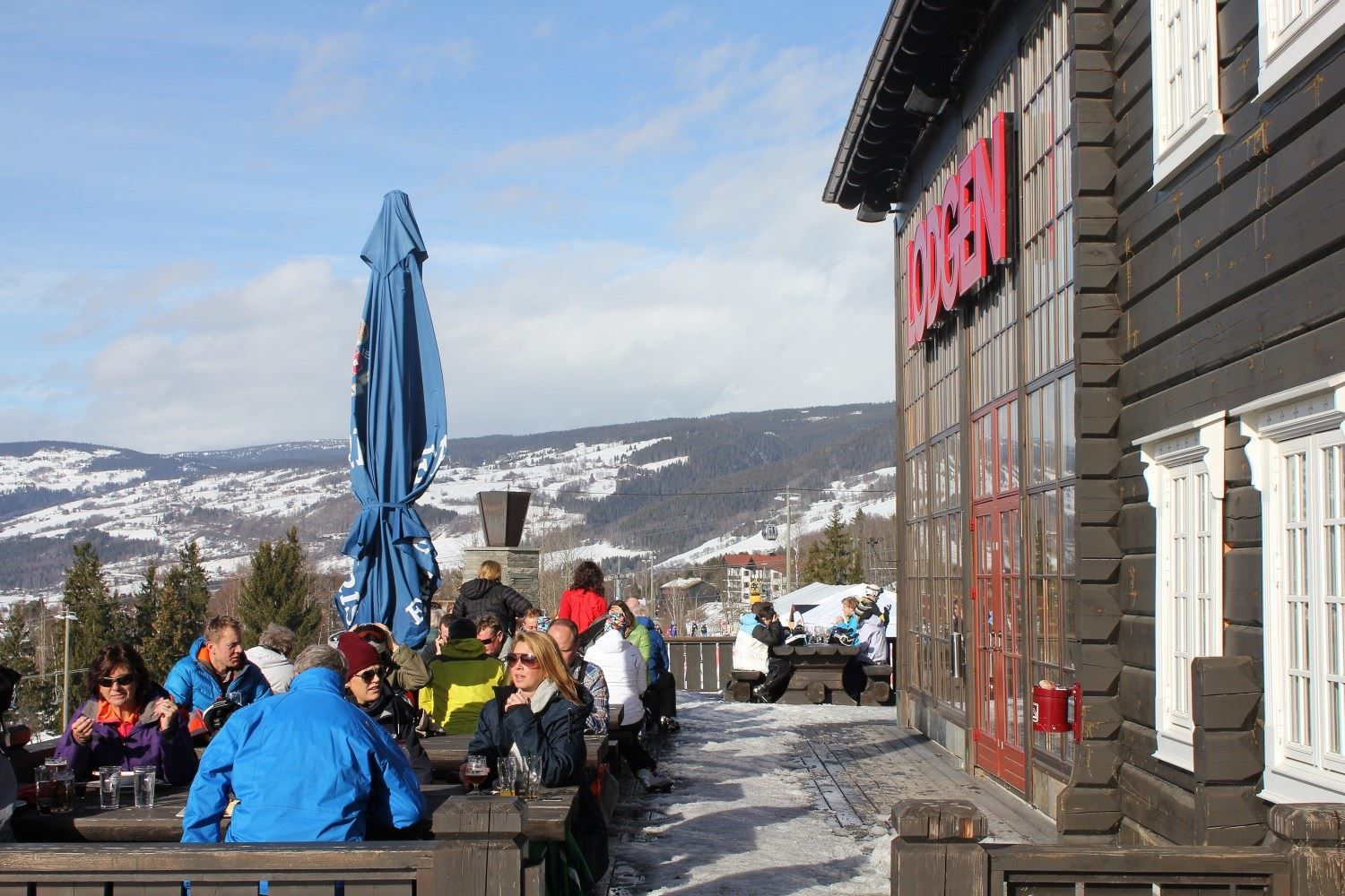Folk sitter på benker utenfor en trebygning med rød skilt, med snødekte fjell og blå himmel i bakgrunnen..