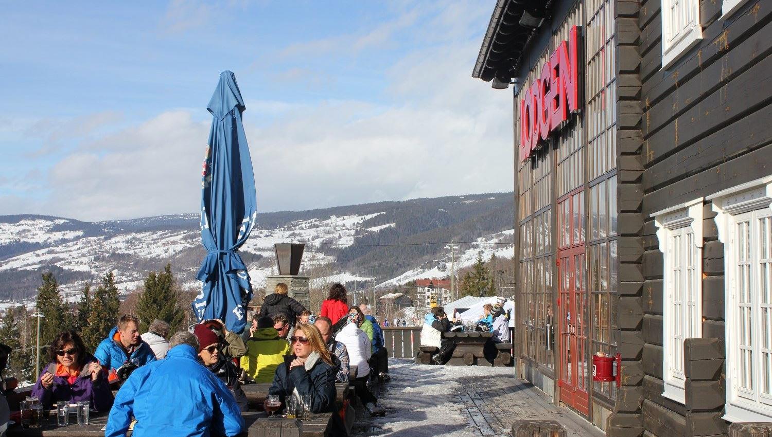 Folk sitter på benker utenfor en trebygning med rød skilt, med snødekte fjell og blå himmel i bakgrunnen..