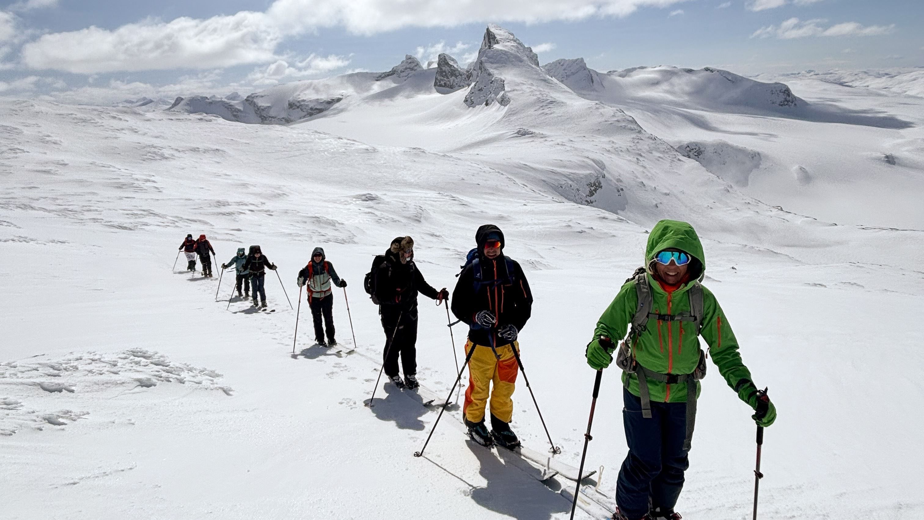 A line of backcountry skiers with poles and backpacks crosses a snowy ridge beneath rugged, cloudlit mountains..