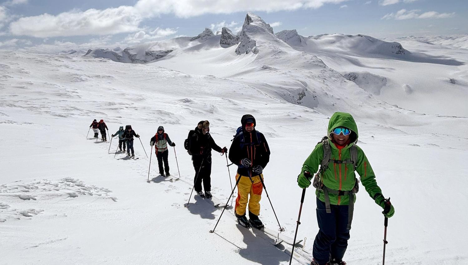 A line of backcountry skiers with poles and backpacks crosses a snowy ridge beneath rugged, cloudlit mountains..