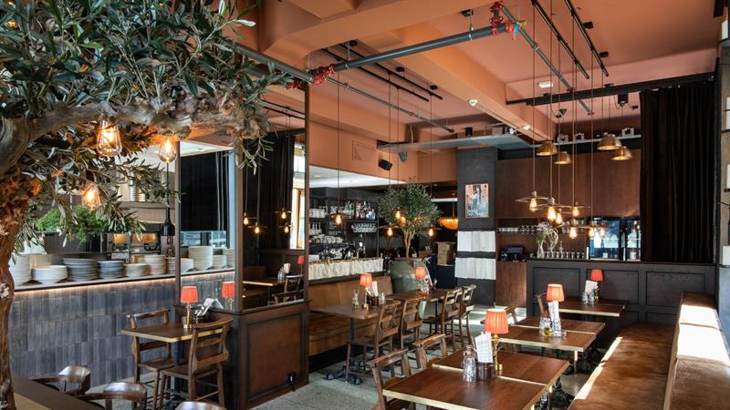 En restaurant med trebord, varme lamper, hengende lyspærer og et stort kunstig oliventre som strekker seg over sitteområdet