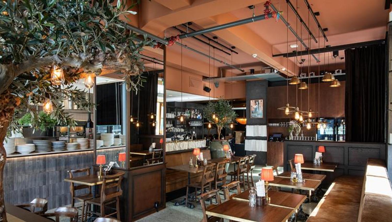 En restaurant med trebord, varme lamper, hengende lyspærer og et stort kunstig oliventre som strekker seg over sitteområdet