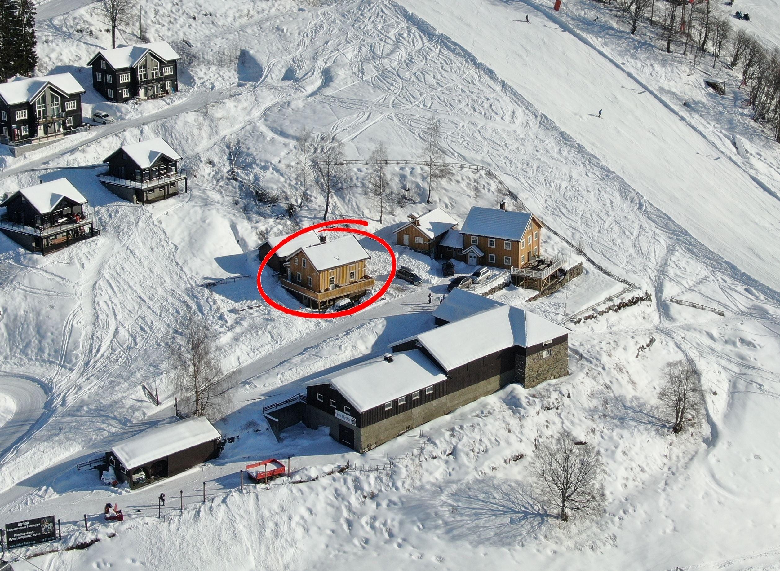 Oversiktsbilde av et snødekt hyttefelt ved en skibakke, med en gul hytte ringet inn i rødt i midten..