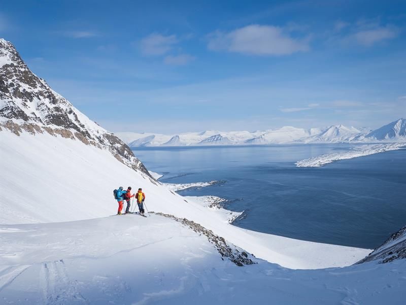 Tre personer står på en fjellhylle å ser ut over fjorden med ski på beina