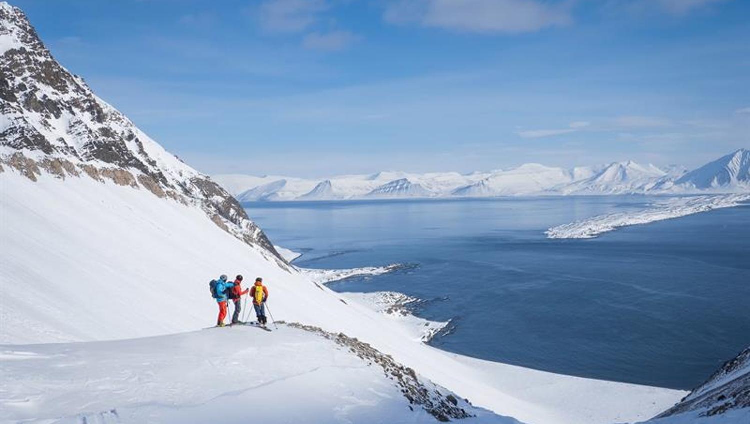 Tre personer står på en fjellhylle å ser ut over fjorden med ski på beina