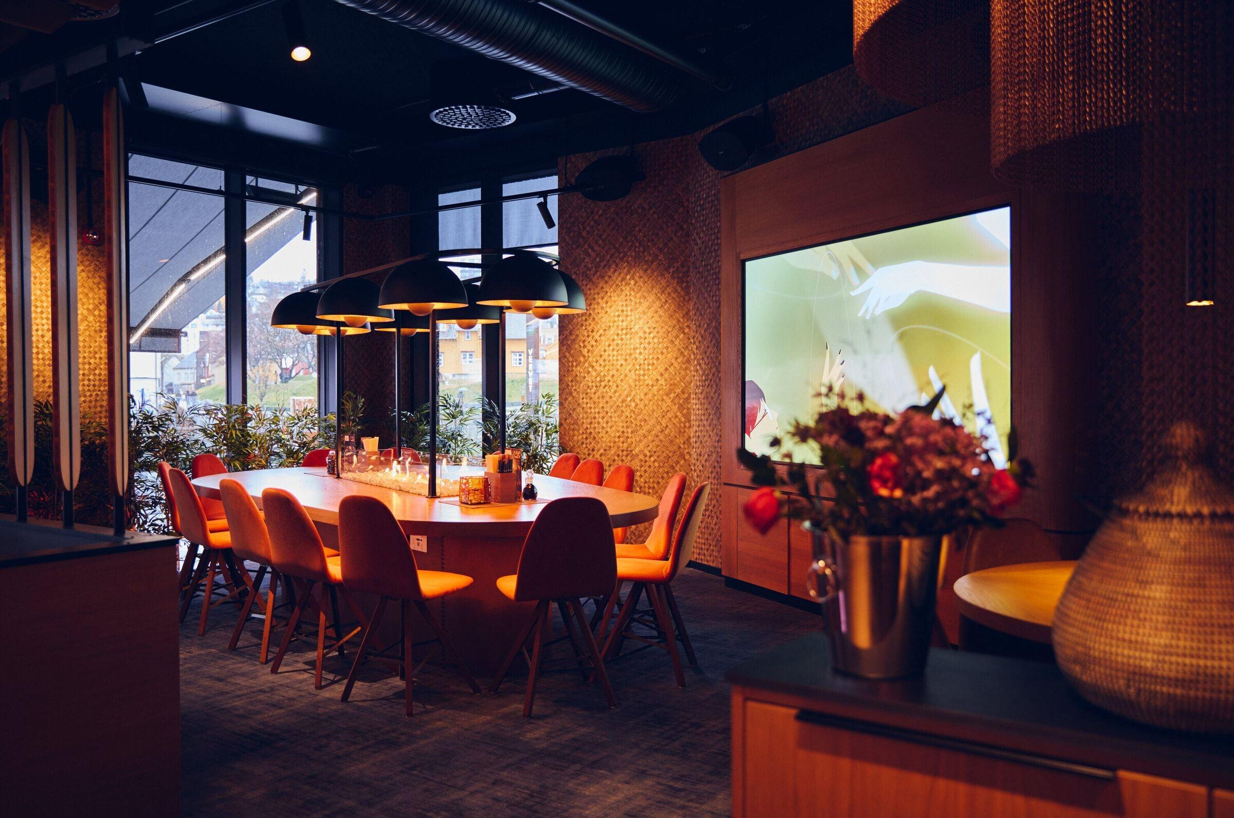 Modern and cosy room with a large oval wooden table, chairs, hanging lamps, and large windows with a view of greenery.