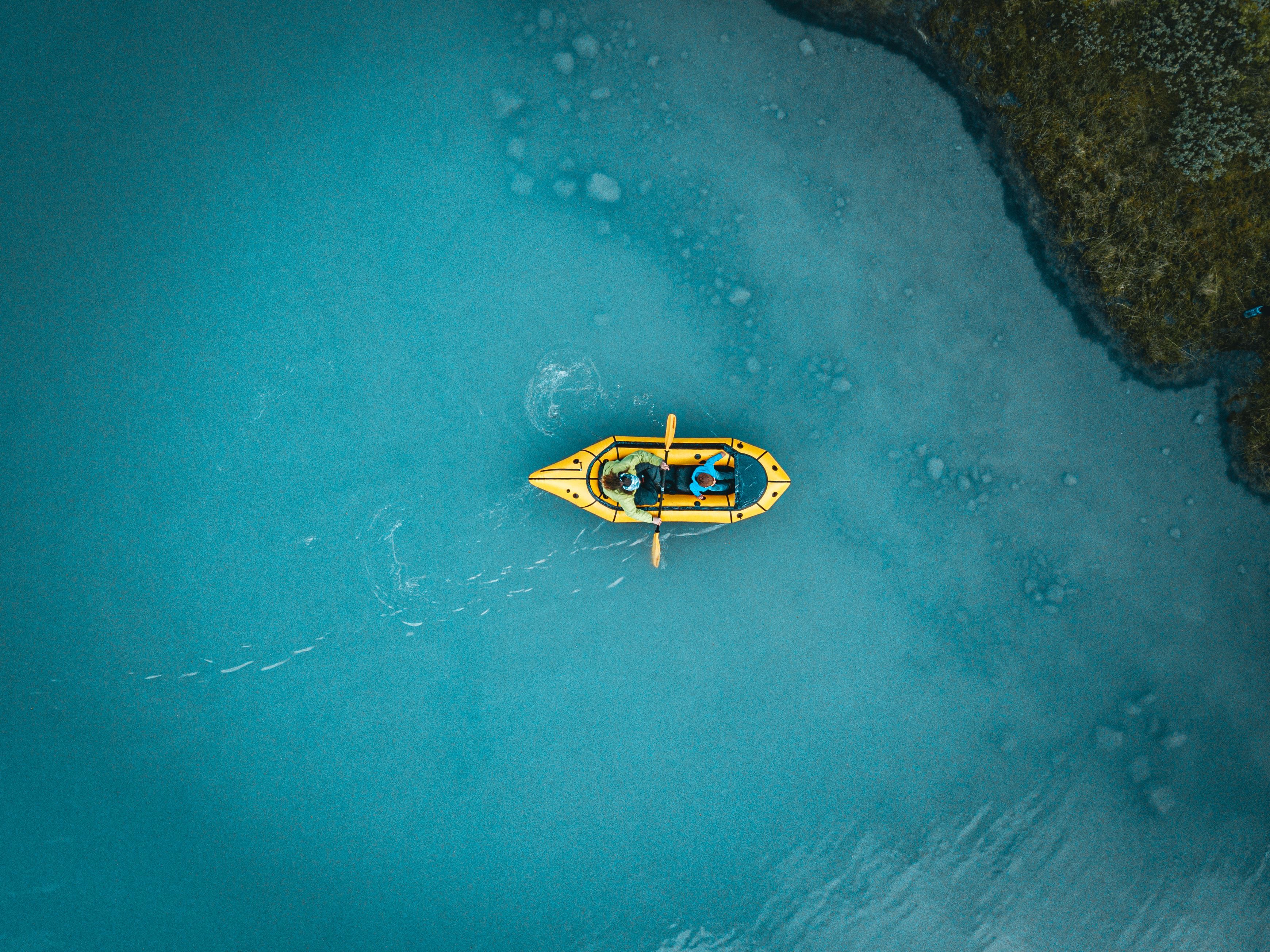 Top-down view of a yellow kayak with two paddlers on calm blue water near a rocky shoreline.