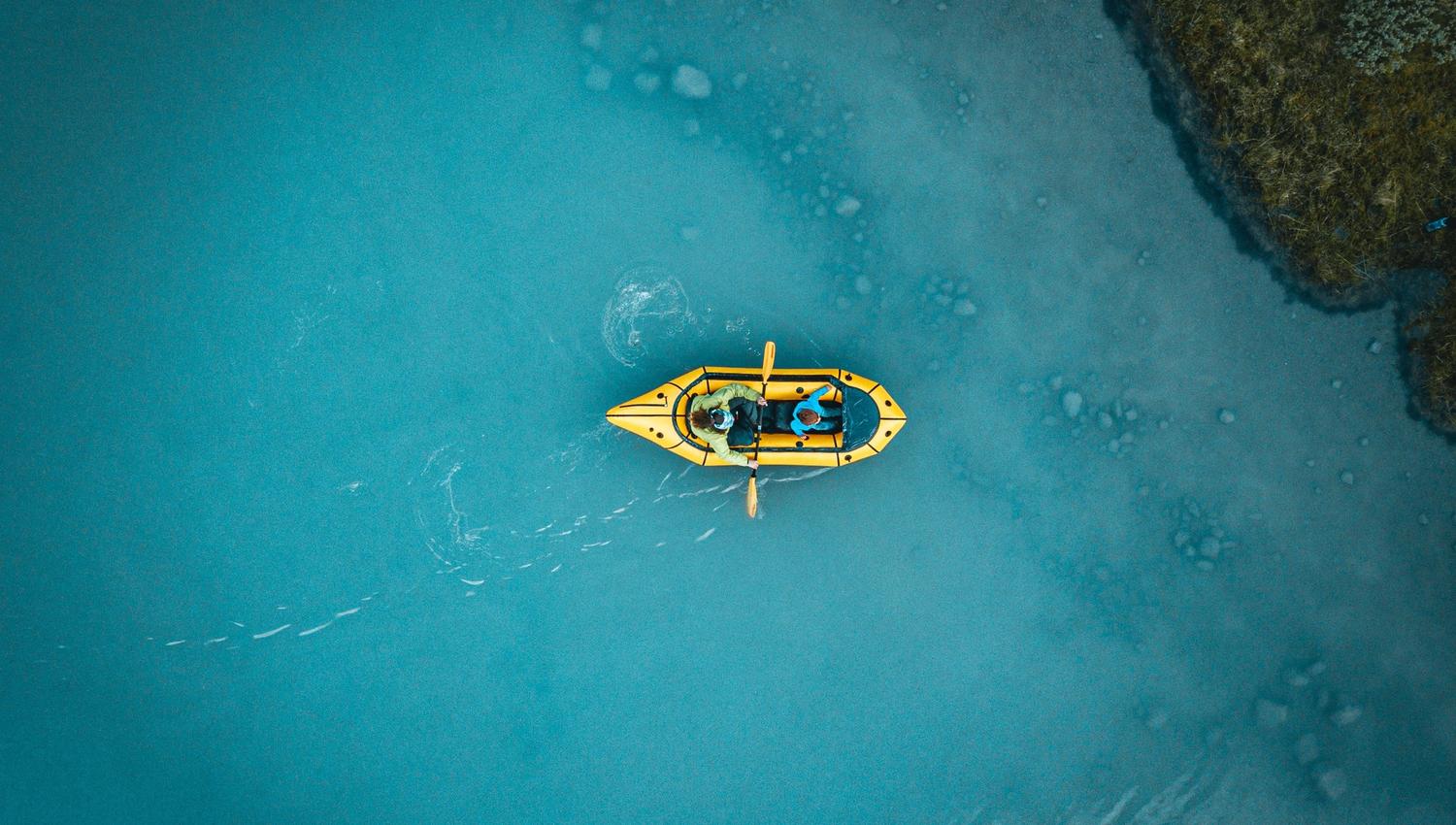 Top-down view of a yellow kayak with two paddlers on calm blue water near a rocky shoreline.