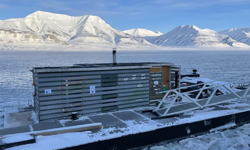 The sauna SvalBad floating in the port of Longyearbyen with snow-covered mountains and a fjord with drift ice in the background
