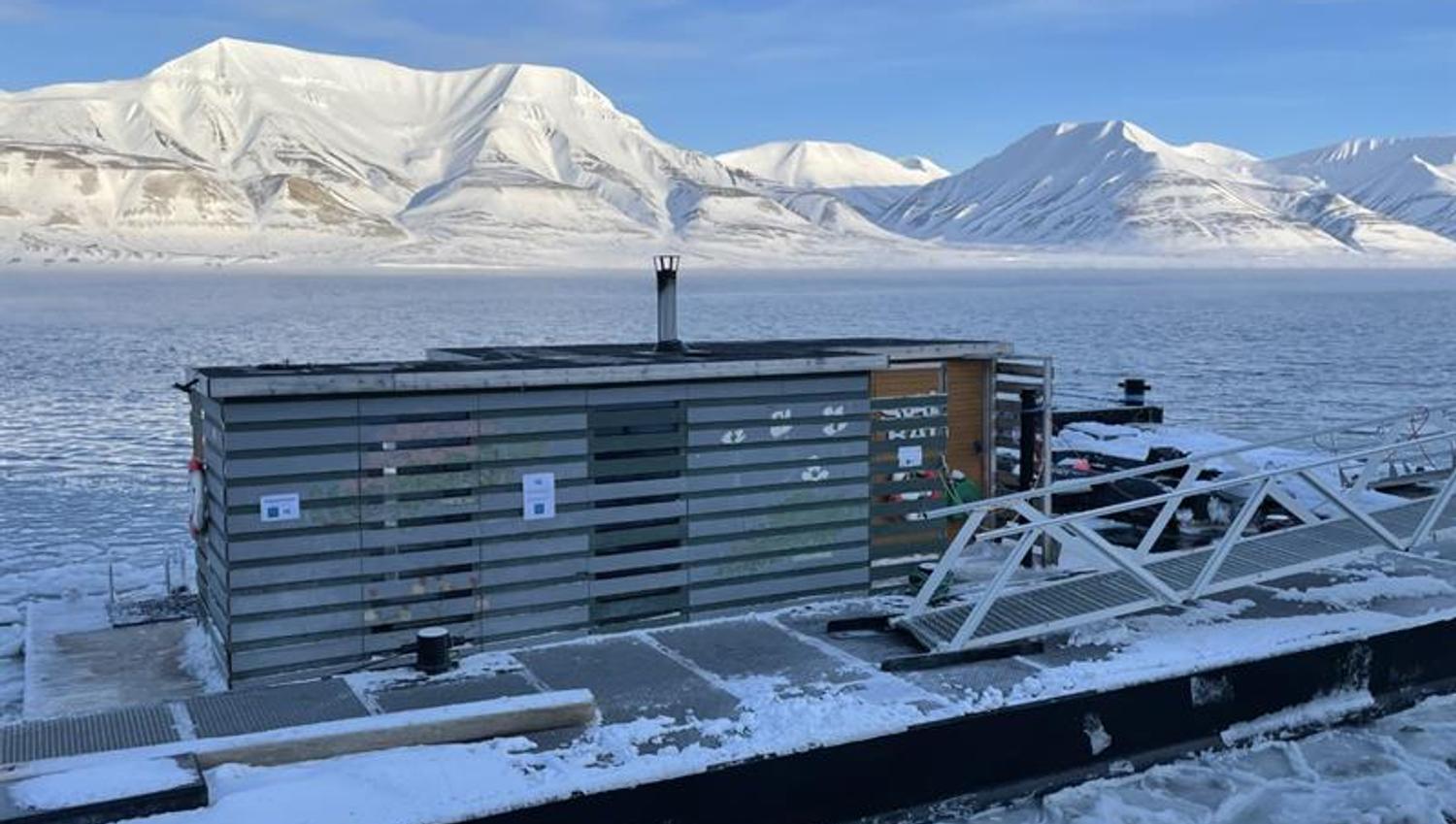 The sauna SvalBad floating in the port of Longyearbyen with snow-covered mountains and a fjord with drift ice in the background