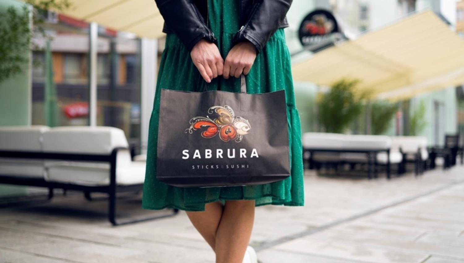 Person i grønn kjole som holder en papirpose med Sabrura logoen