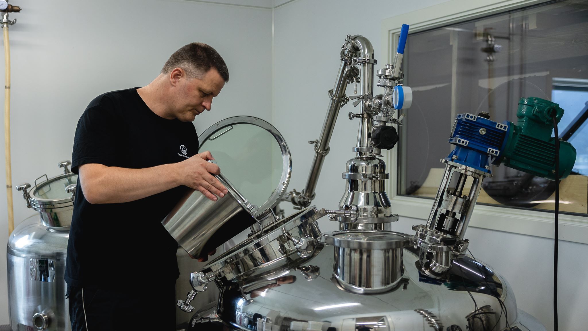 Person in a black shirt pours liquid from a metal container into an open stainless-steel industrial tank indoors.