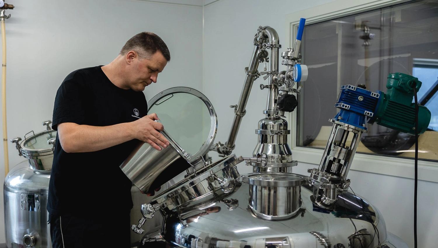 Person in a black shirt pours liquid from a metal container into an open stainless-steel industrial tank indoors.