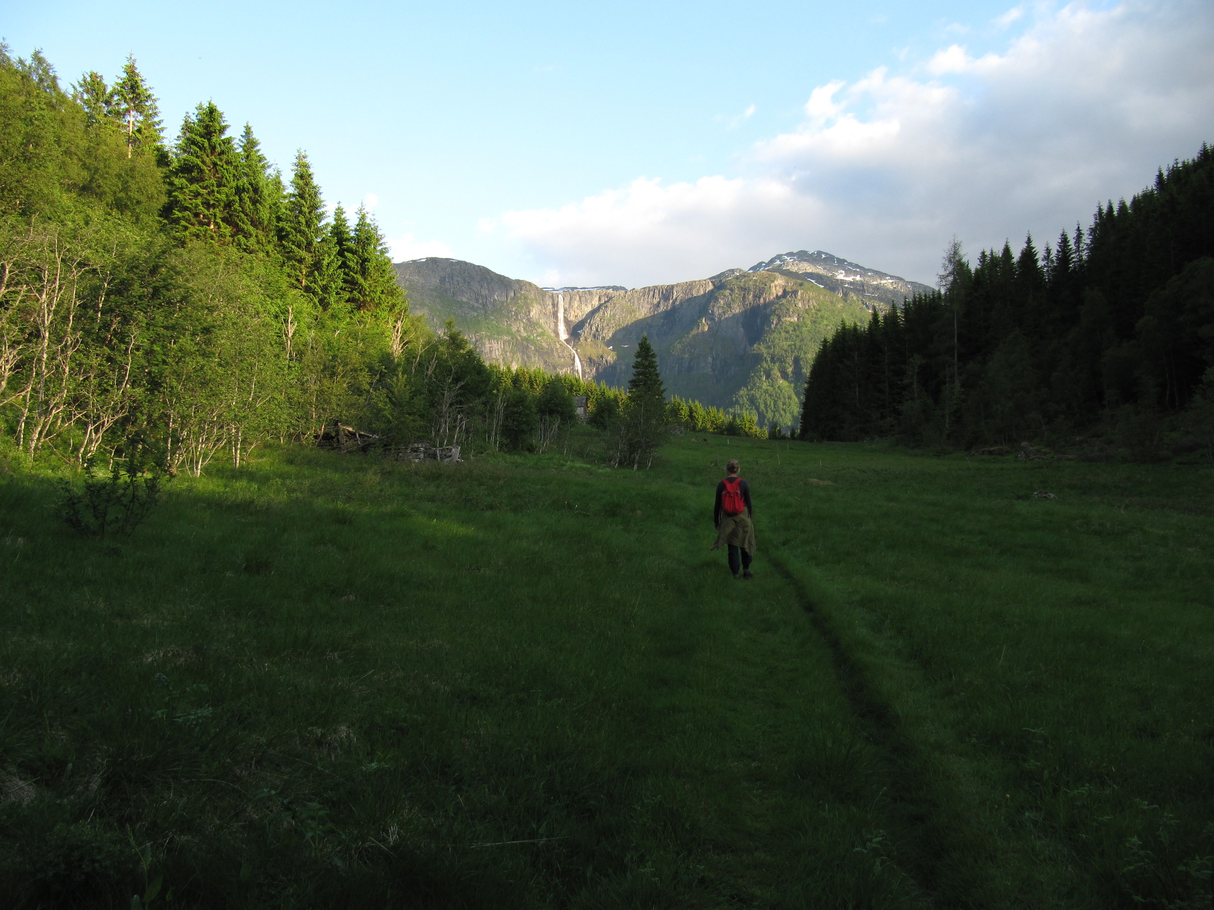 Hiker with red backpack walking through the valley on the Hovden rundt trail, with mountains and a distant waterfall.