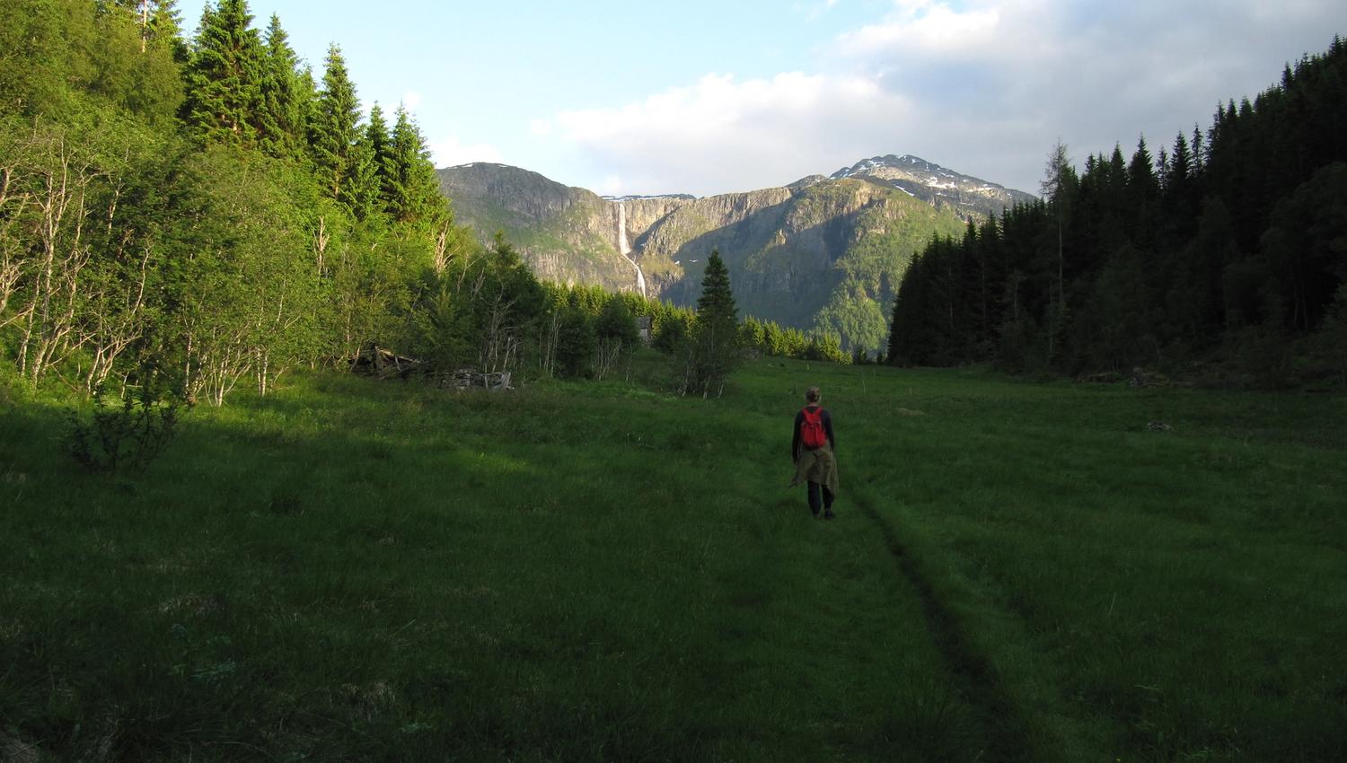 Hiker with red backpack walking through the valley on the Hovden rundt trail, with mountains and a distant waterfall.