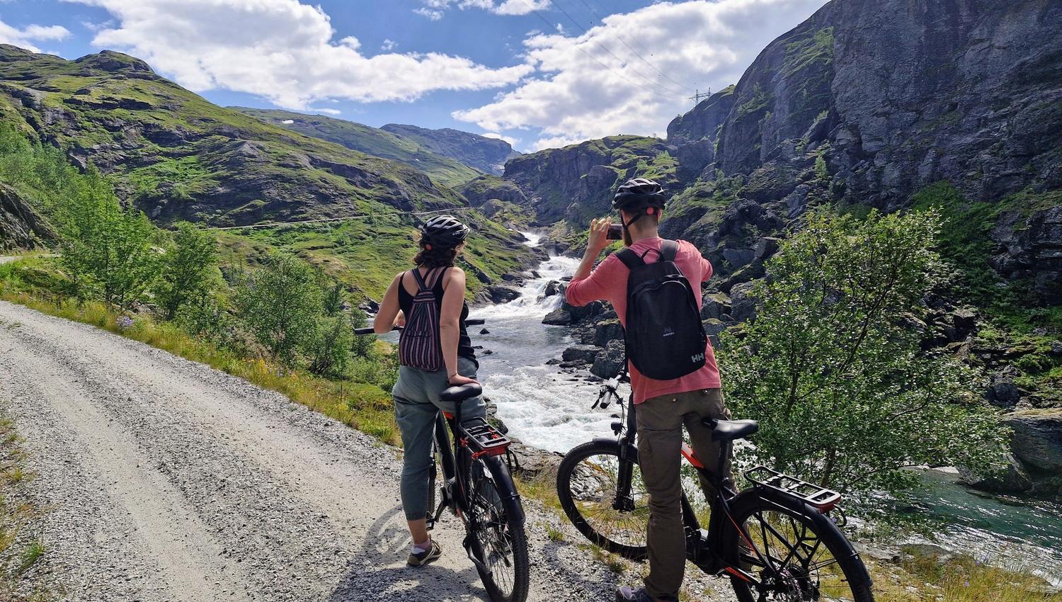 Eit par har stoppa for å ta bilete av ei elv, langs Rallarvegen. Dei står med kvar sin sykkel, på ein fin sommardag.