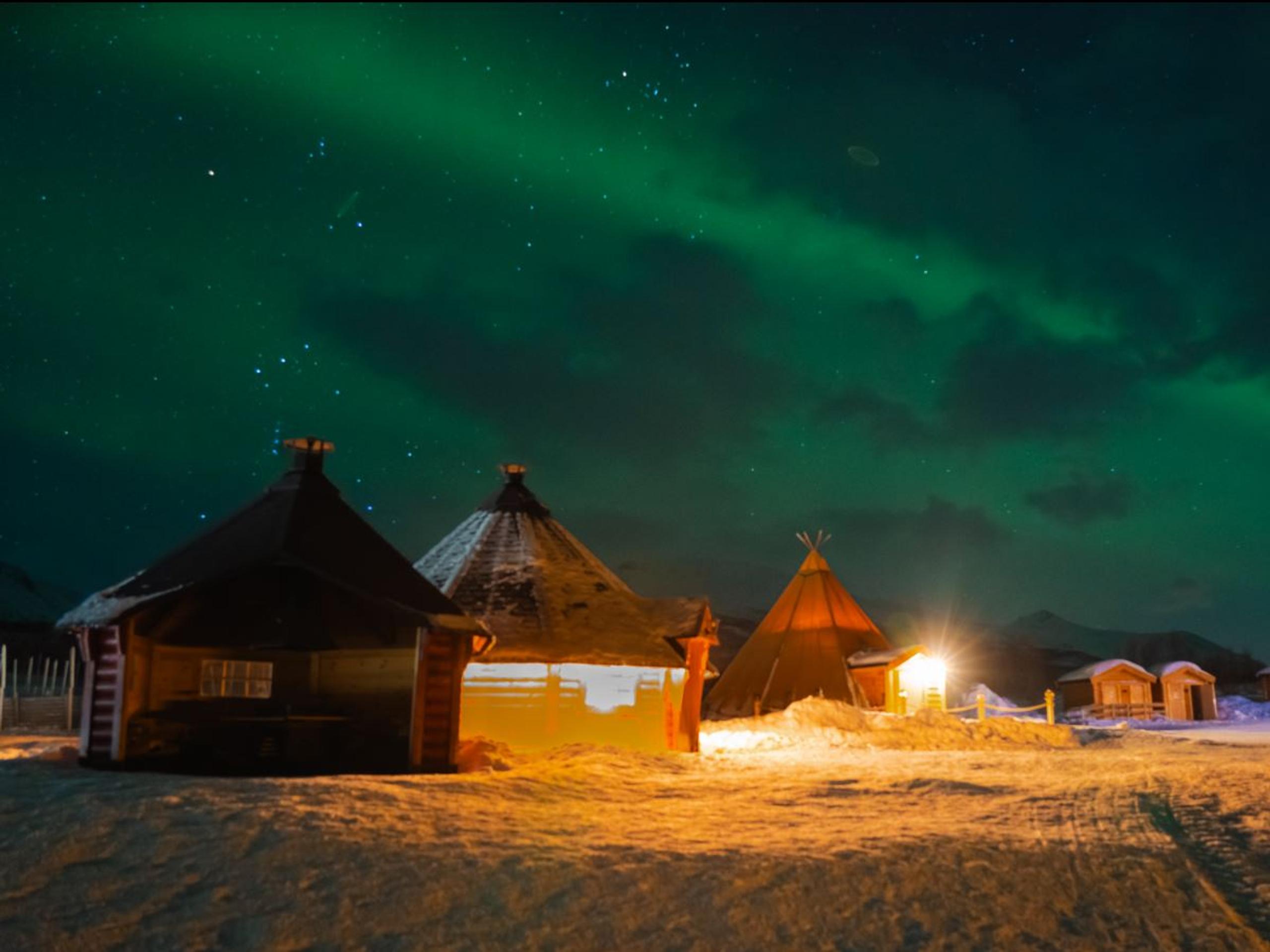 Northern Lights above snow-covered lavvus and buildings at Tromsø Lapland’s Sámi camp near Tromsø.