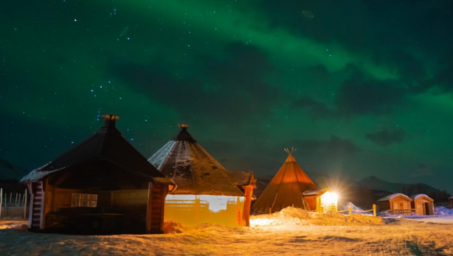 Nordlys over snødekte hytter og lavvo opplyst av varme lys.