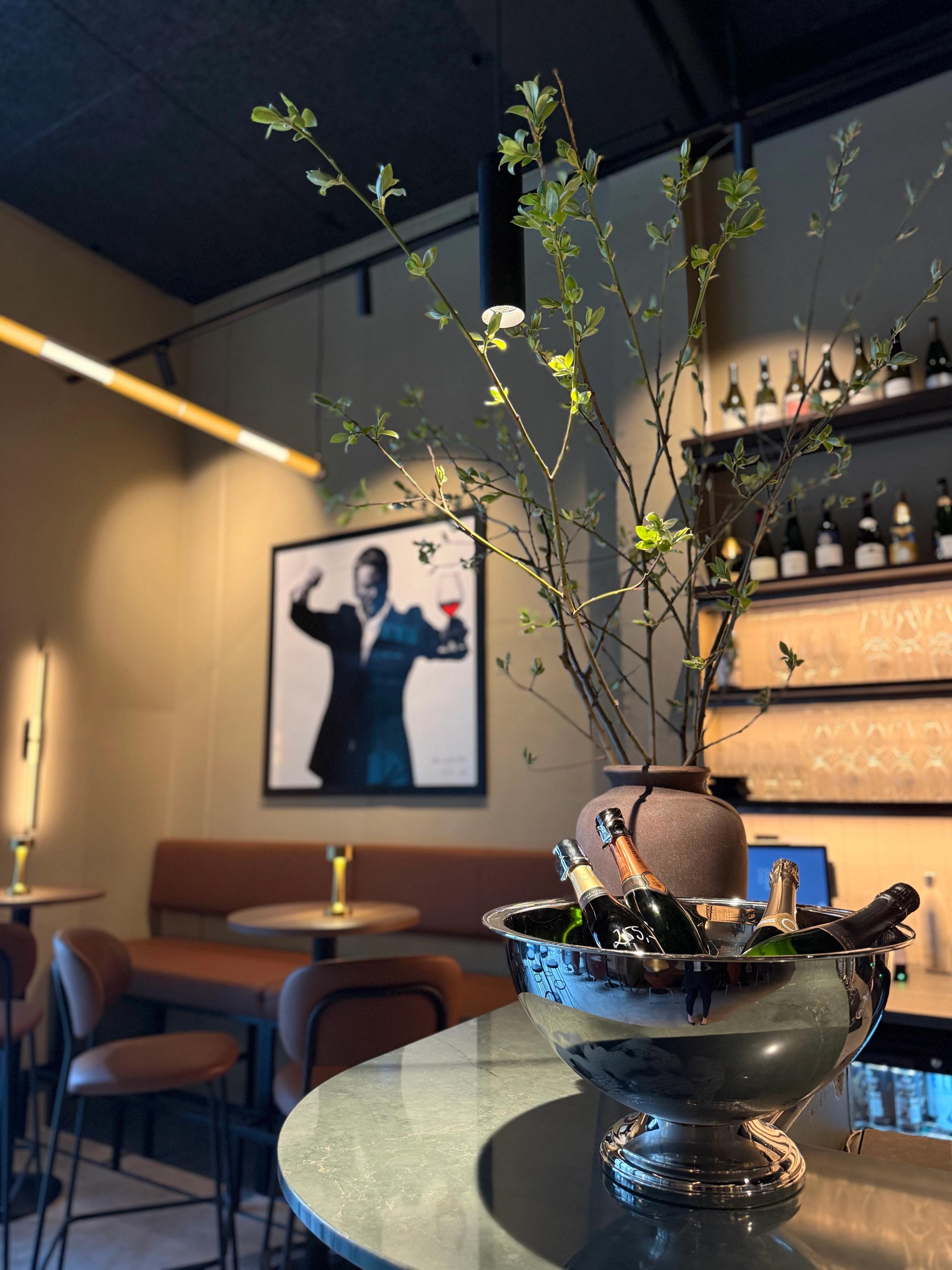 Metal ice bucket with champagne bottles and tall green branches on a bar counter, with lounge seating and bottle shelves behind.
