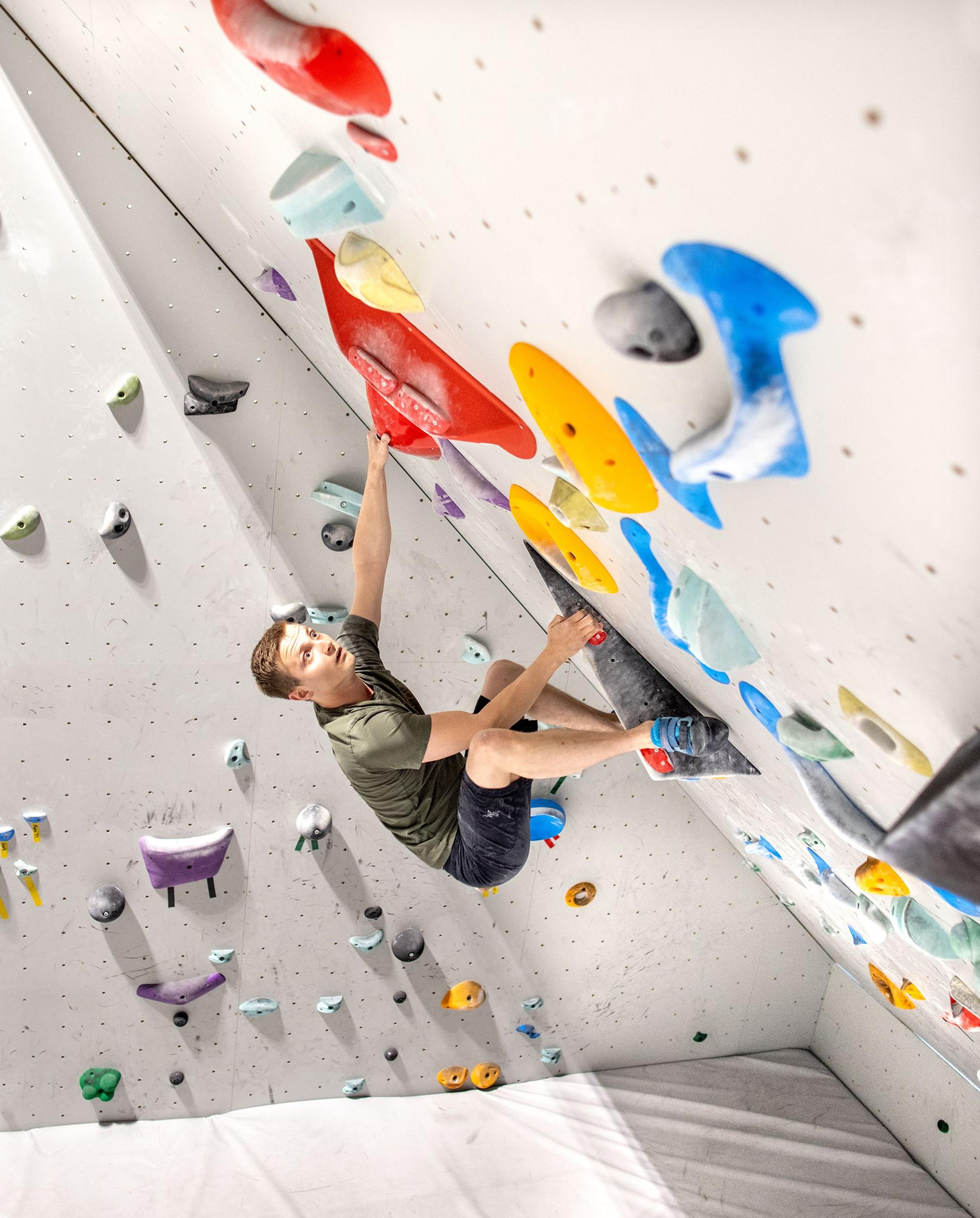 Man bouldering on an overhanging indoor climbing wall, reaching for a large red hold among colorful grips.