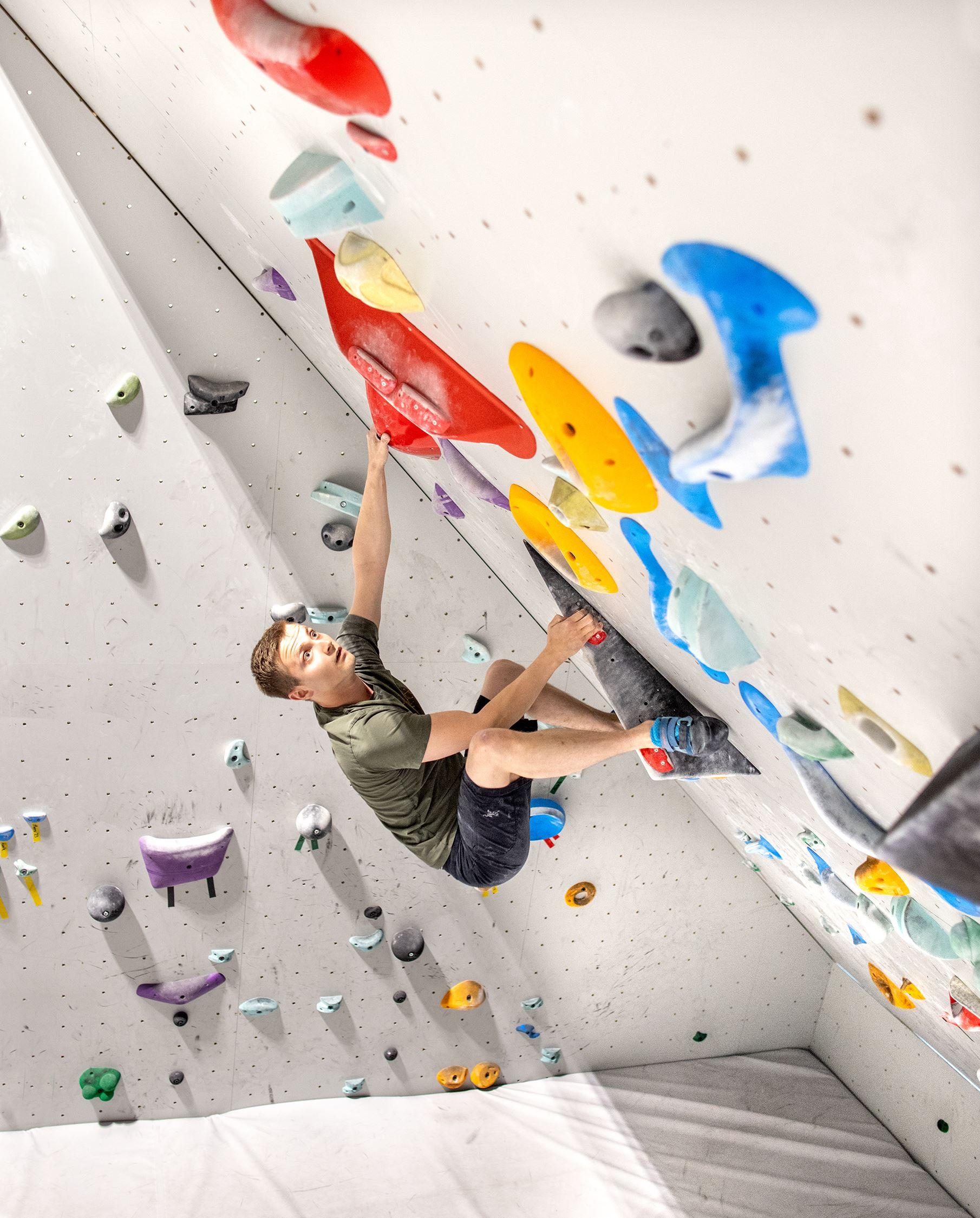 Man bouldering on an overhanging indoor climbing wall, reaching for a large red hold among colorful grips.