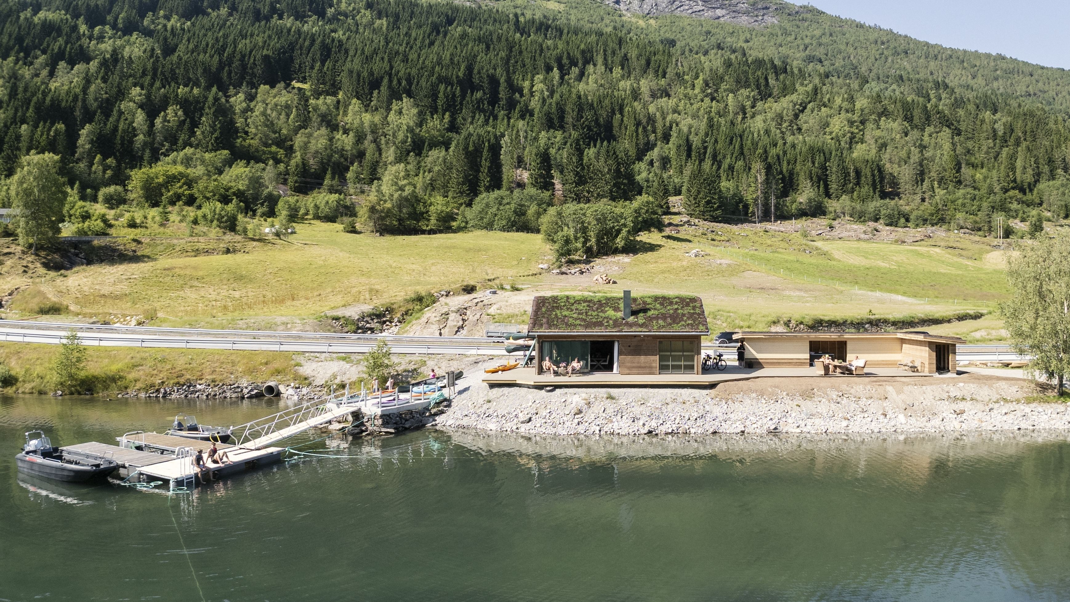 Modern wooden cabin with a green roof beside a calm lake, with a dock, boats, and forested hills behind..
