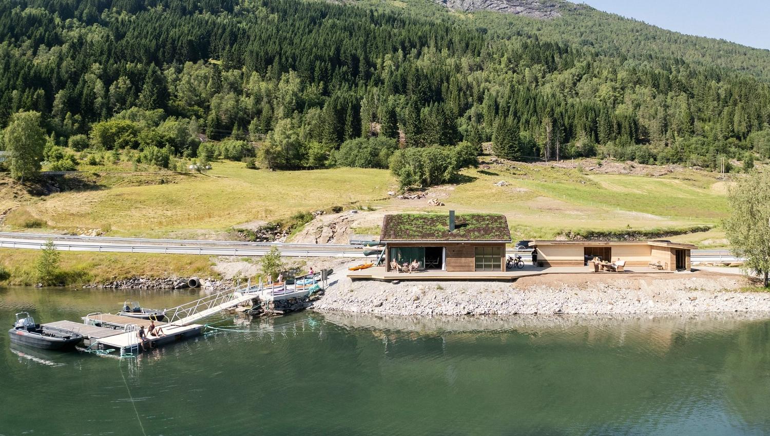 Modern wooden cabin with a green roof beside a calm lake, with a dock, boats, and forested hills behind..
