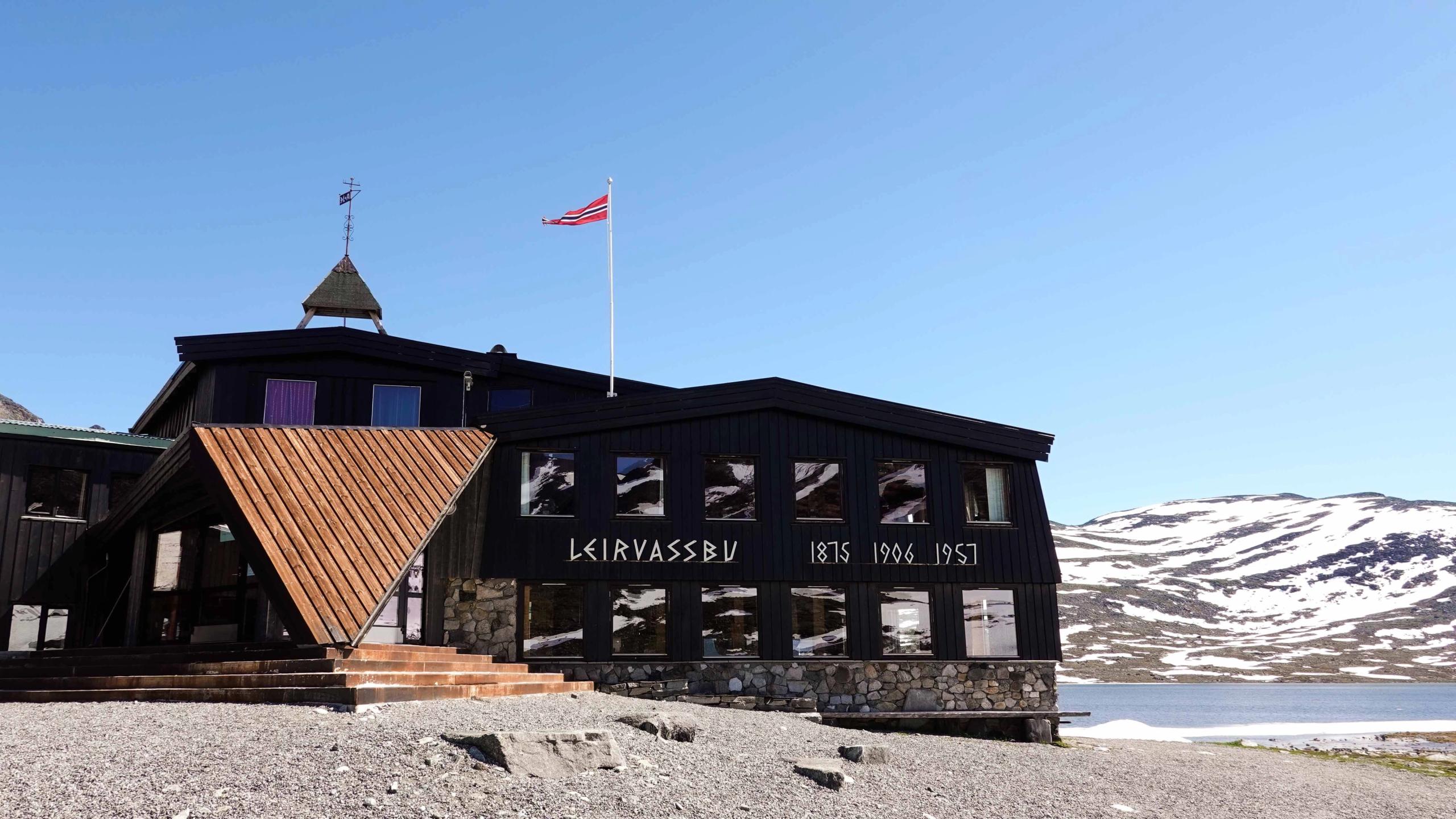 Svart fjellbygning med steinsokkel og trekledd inngang, flaggstong på taket, ved vatn og snødekte fjell..