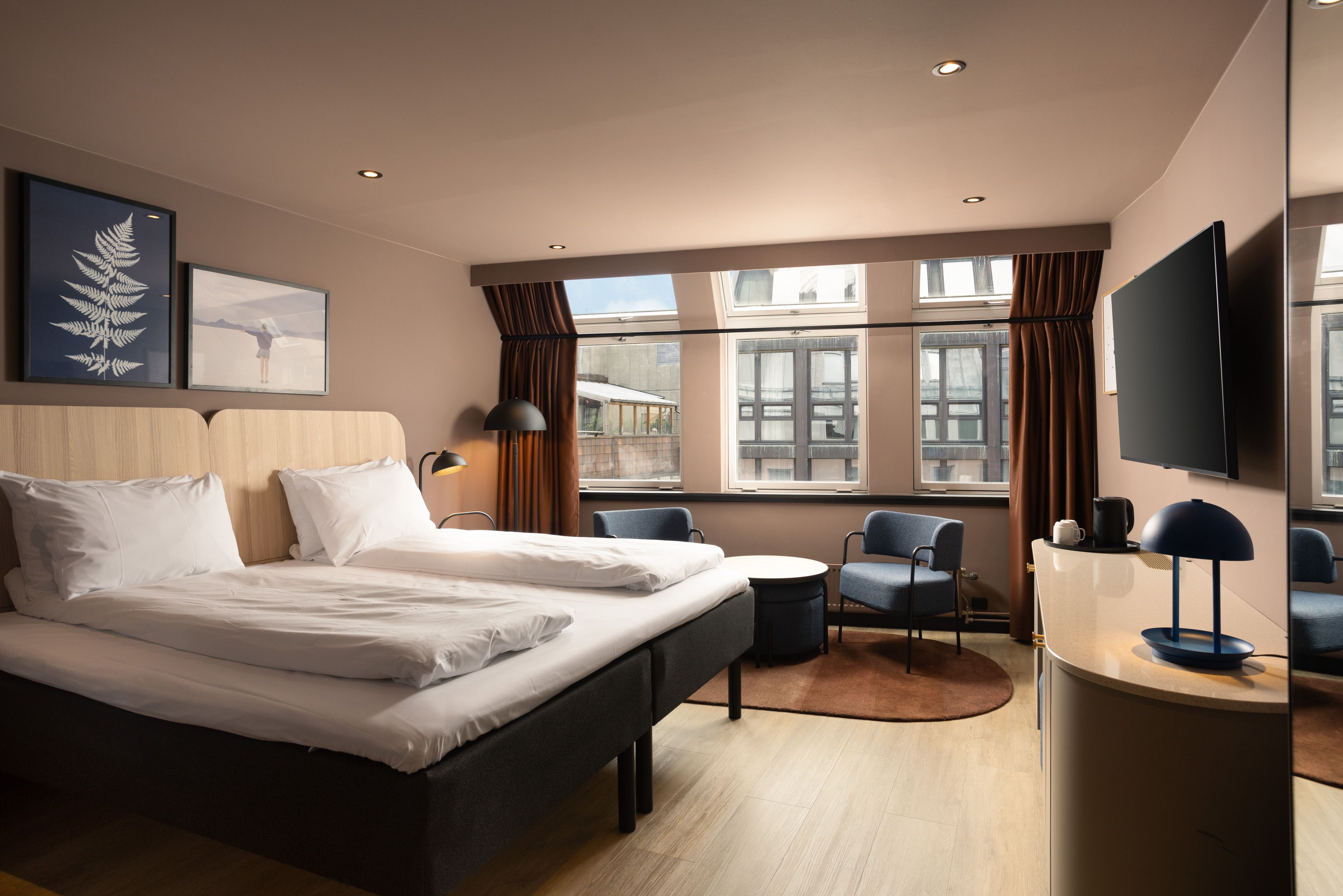 Hotel room with large double bed, TV on the wall, blue armchairs and brown curtains