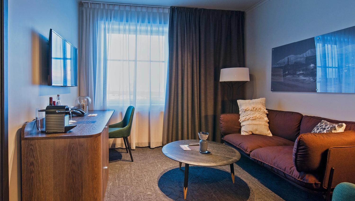 A hotel room with a sofa, coffee table and work desk