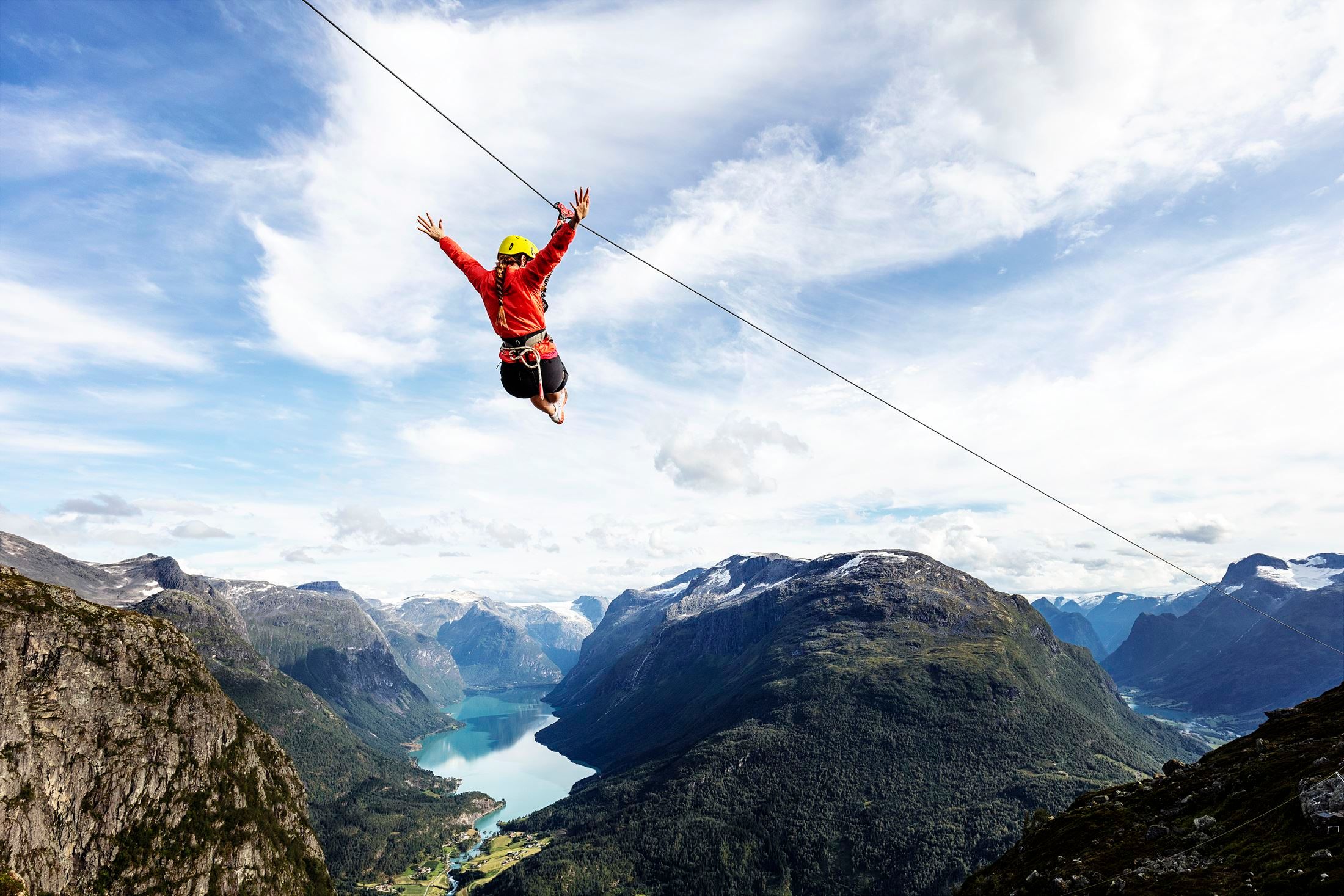 En person henger i en line over fjell og en lang fjord under skyet blå himmel..