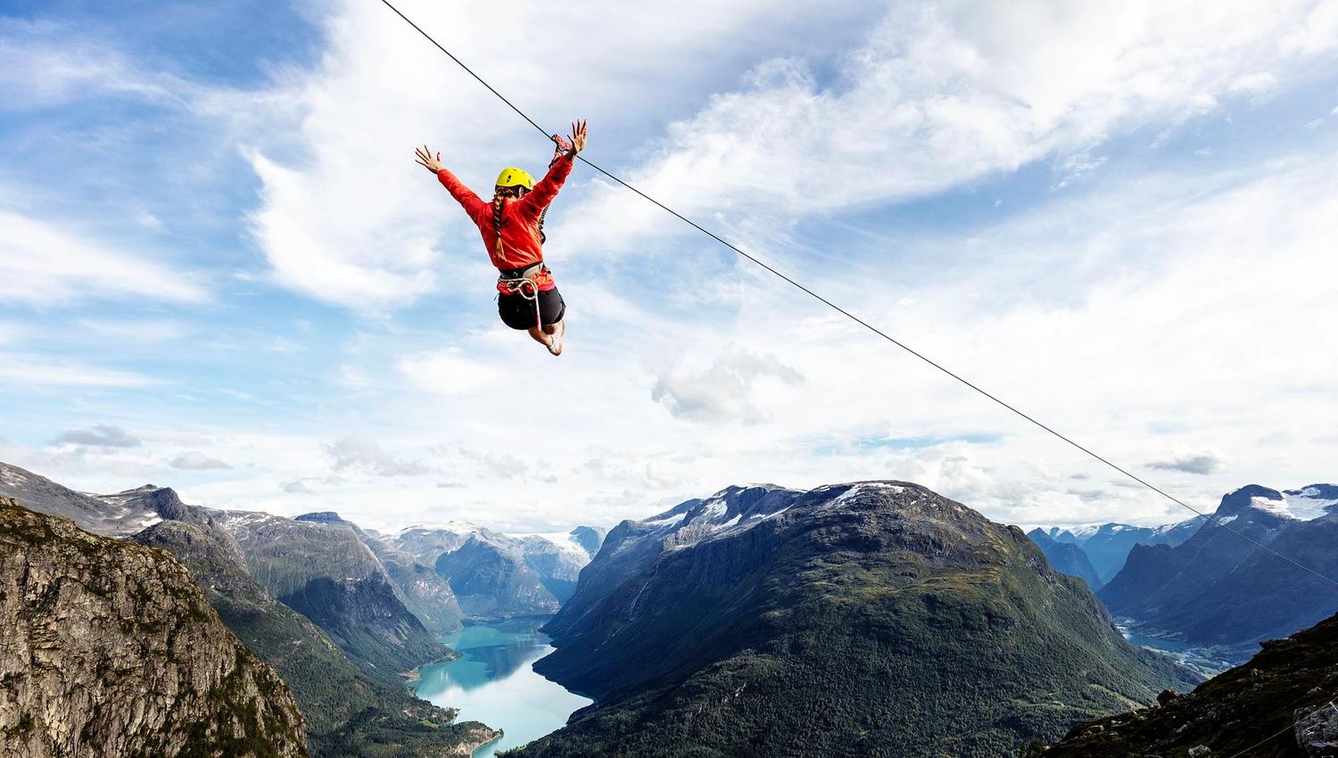 En person henger i en line over fjell og en lang fjord under skyet blå himmel..
