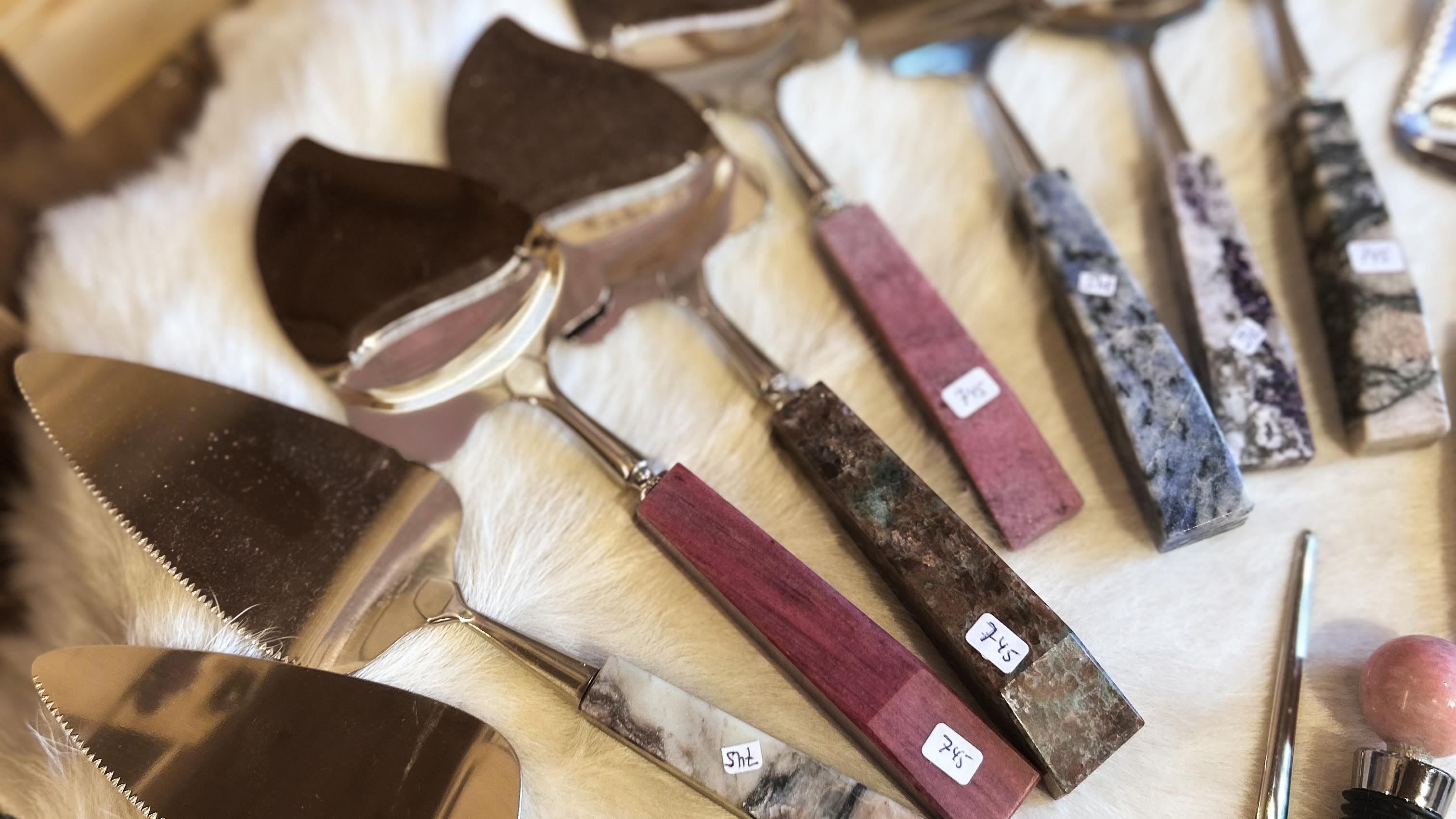 Close-up of assorted metal cheese knives and slicers with colorful stone and wood handles on white fur..