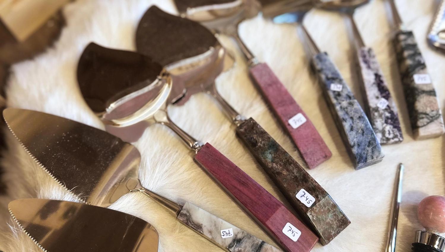 Close-up of assorted metal cheese knives and slicers with colorful stone and wood handles on white fur..