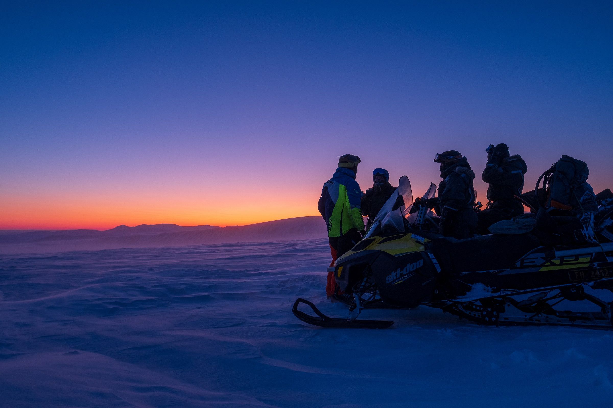 A group of guests and a guide standing behind a snowmobile looking out across a landscape with blue and orange twillight colours in the horizon