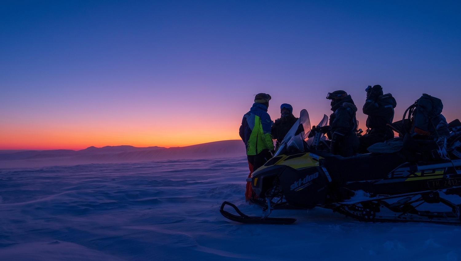 A group of guests and a guide standing behind a snowmobile looking out across a landscape with blue and orange twillight colours in the horizon