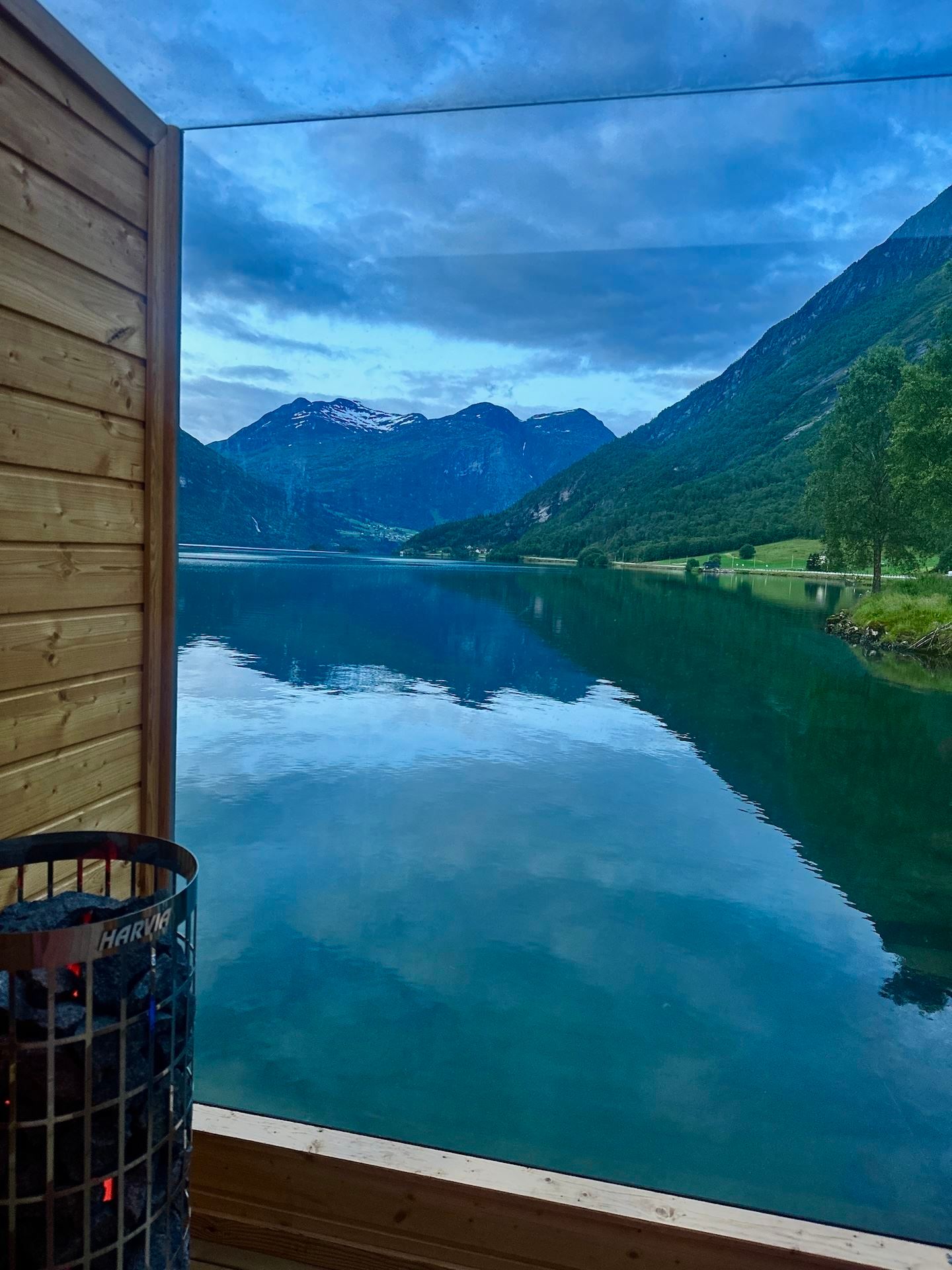 View through a glass wall from a wooden sauna, overlooking a calm lake, green mountains, and cloudy sky..