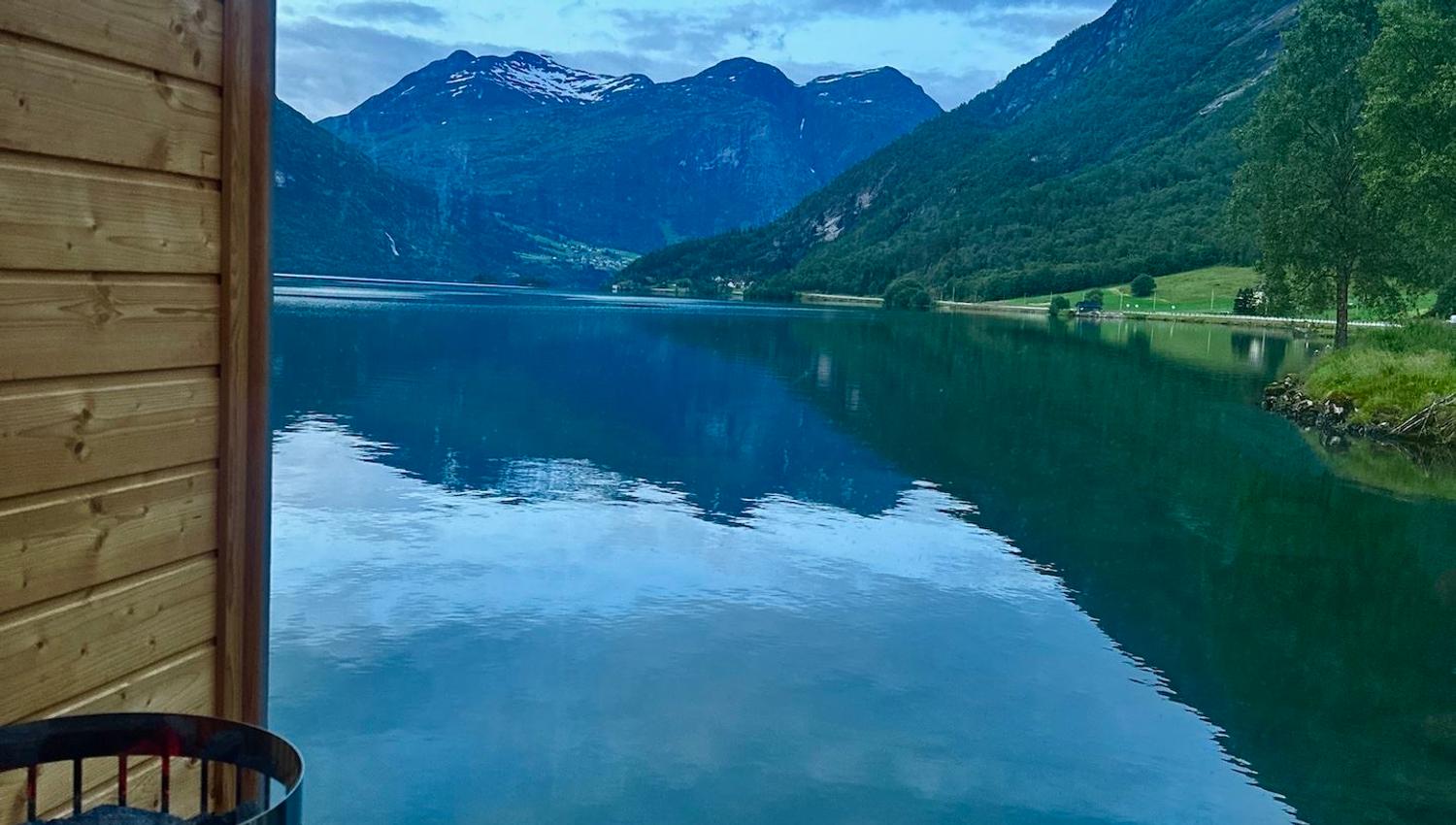 View through a glass wall from a wooden sauna, overlooking a calm lake, green mountains, and cloudy sky..