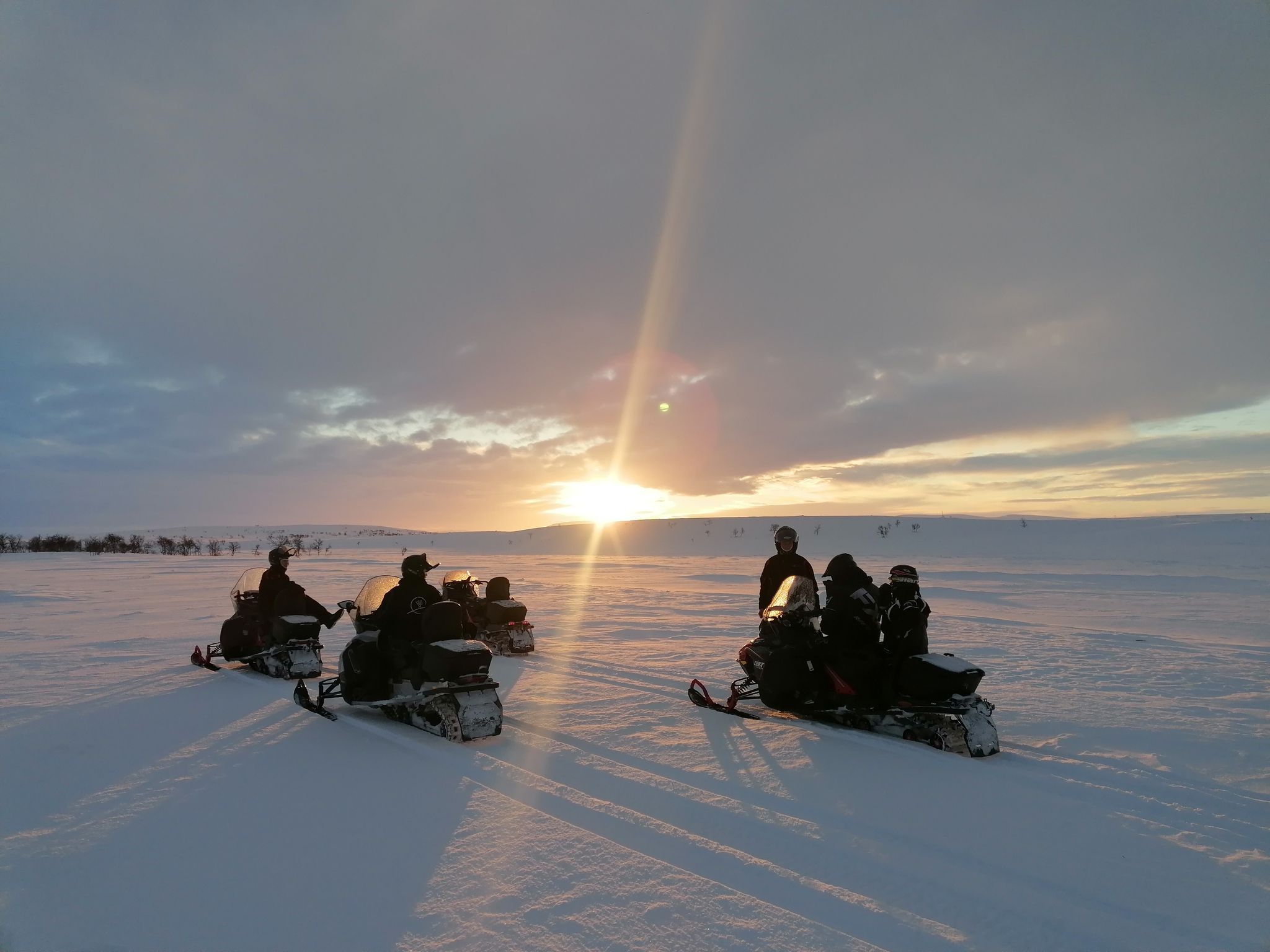 Quatre personnes sur des motoneiges, admirant le coucher de soleil sur un paysage enneigé.