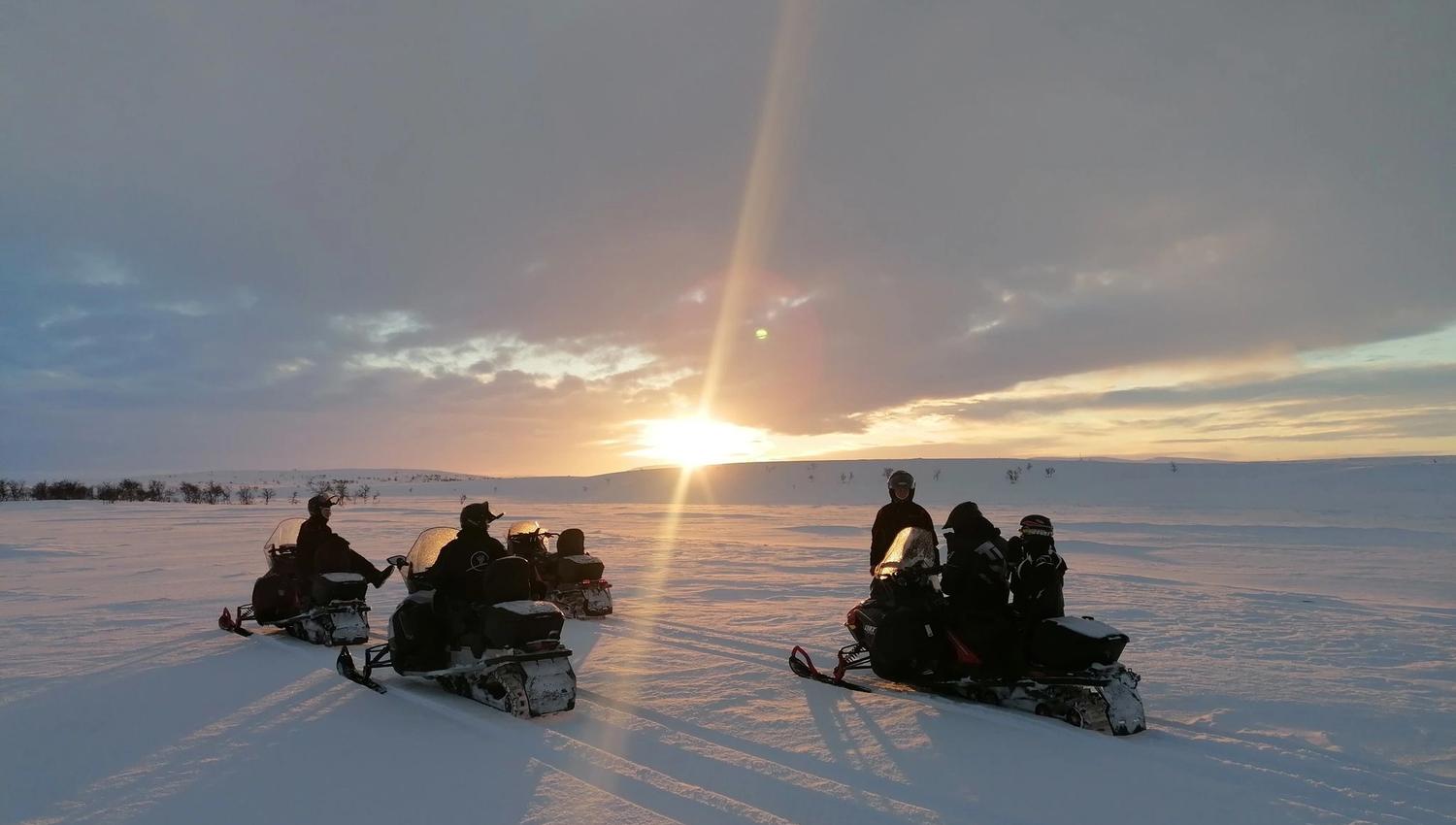 Quatre personnes sur des motoneiges, admirant le coucher de soleil sur un paysage enneigé.