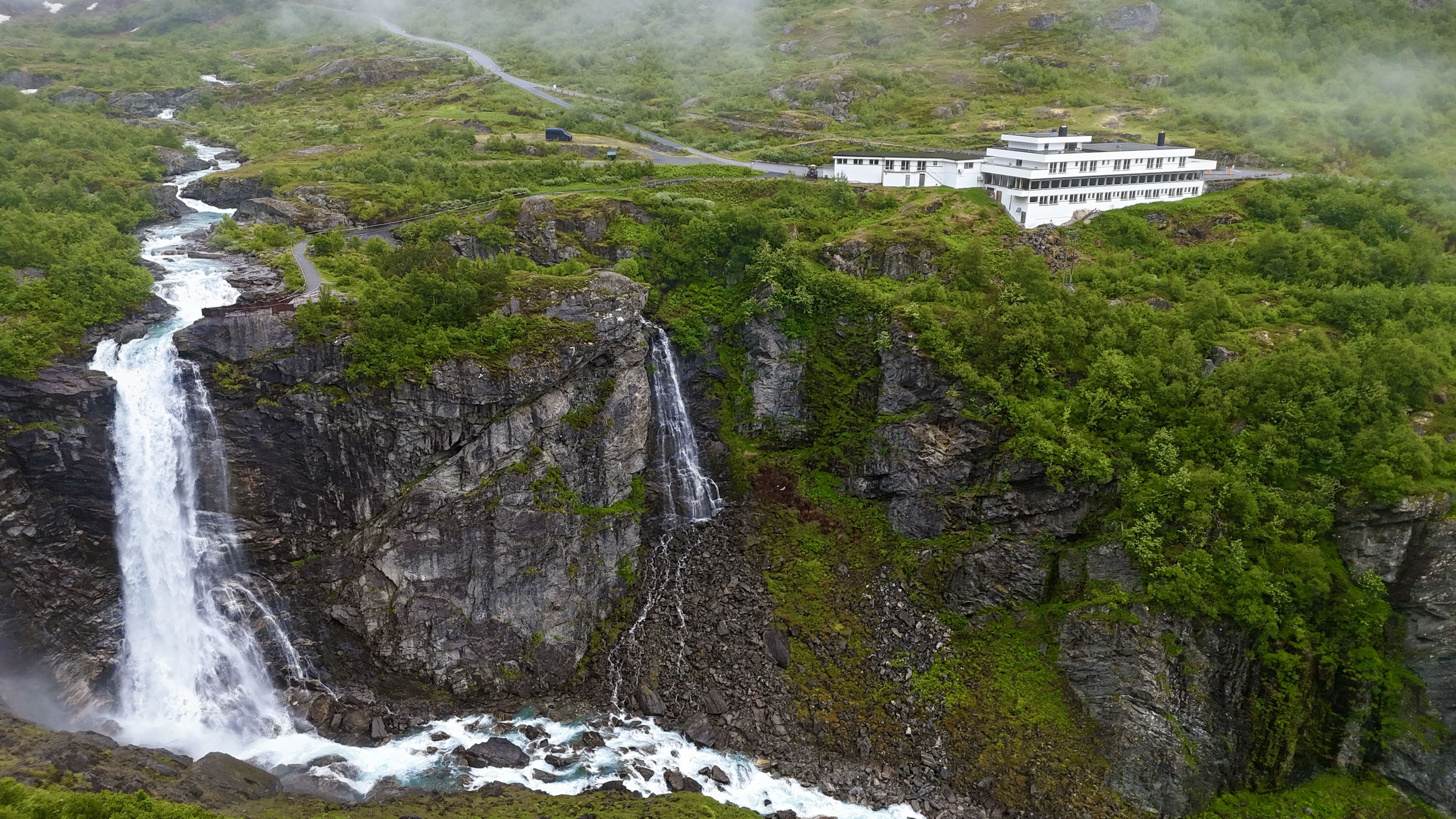 Aerial view of a white hotel on a green rocky cliff above twin waterfalls feeding a foaming river in mist..