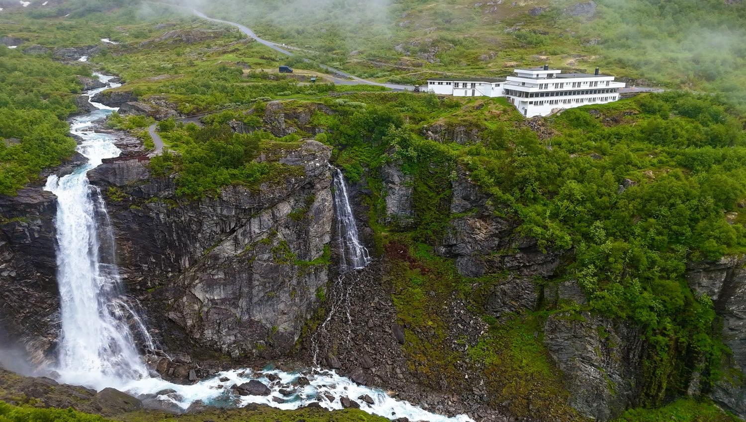 Aerial view of a white hotel on a green rocky cliff above twin waterfalls feeding a foaming river in mist..