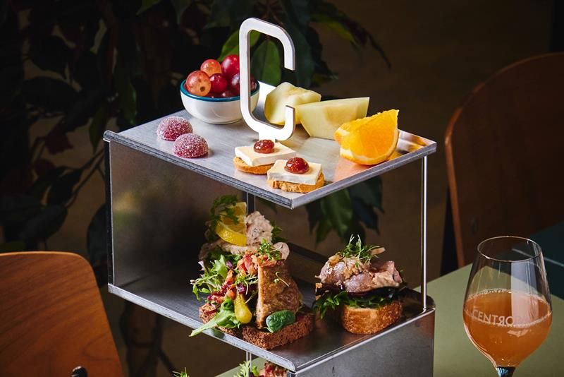 A modern three-tiered metal stand displaying a selection of small open-faced sandwiches, cheeses with jam, fruit slices, grapes, and sweets. A glass o