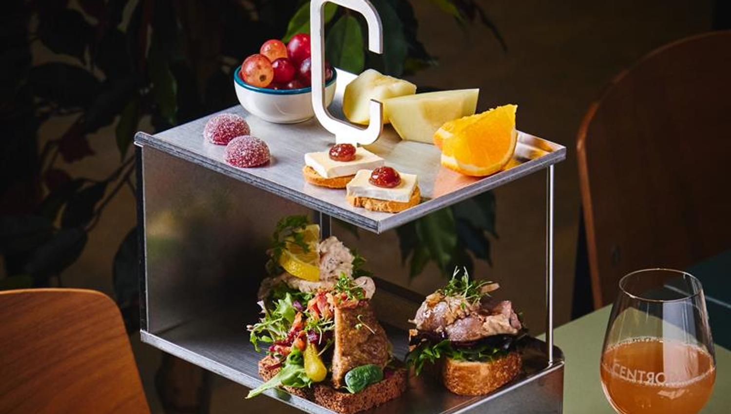 A modern three-tiered metal stand displaying a selection of small open-faced sandwiches, cheeses with jam, fruit slices, grapes, and sweets. A glass o