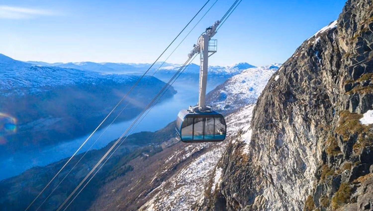 A cable car gondola hangs from wires beside a steep rocky cliff, overlooking a snowy mountain valley and fjord.