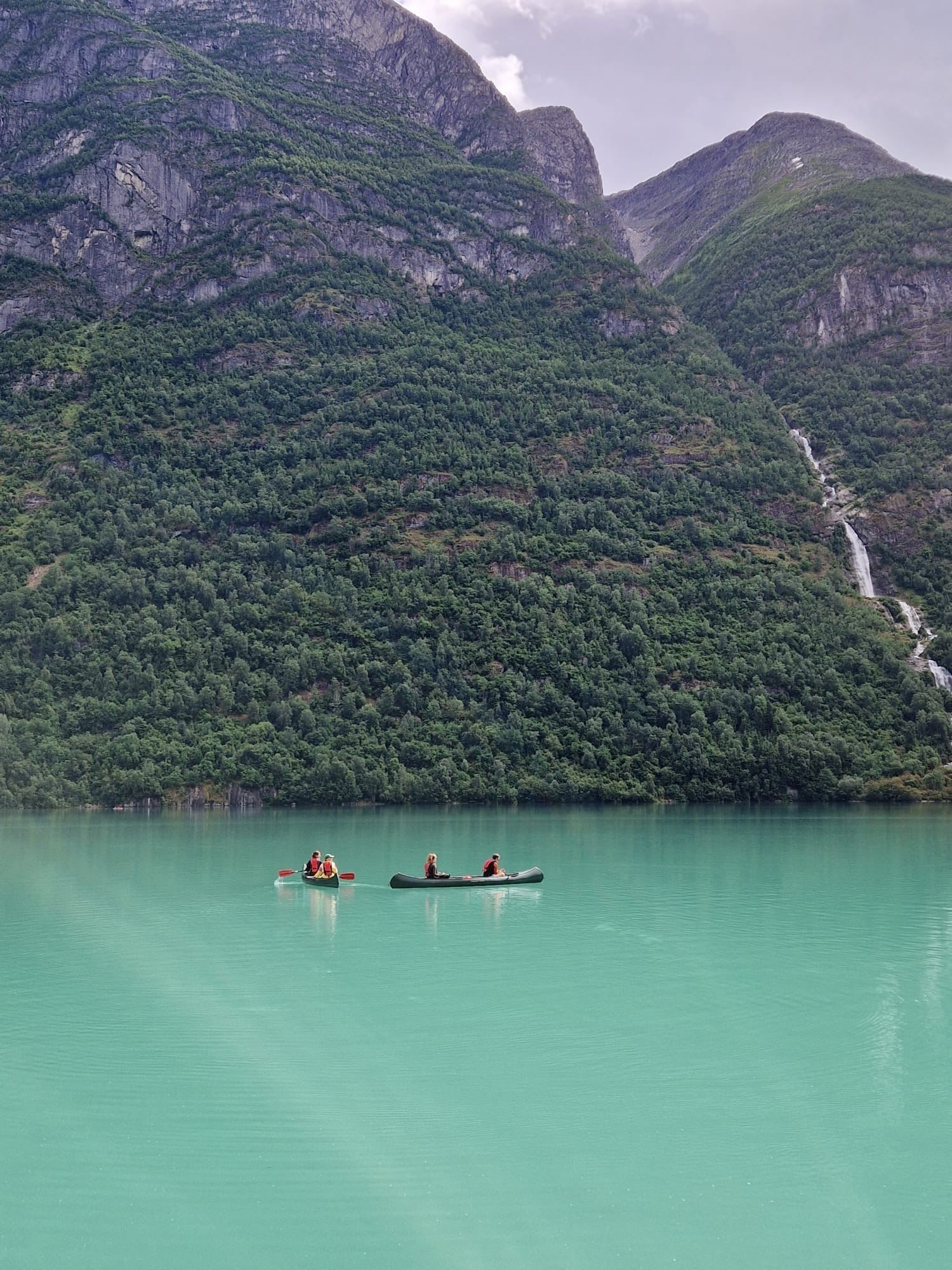 Two canoes with paddlers float on calm turquoise water below forested mountains and a distant waterfall..