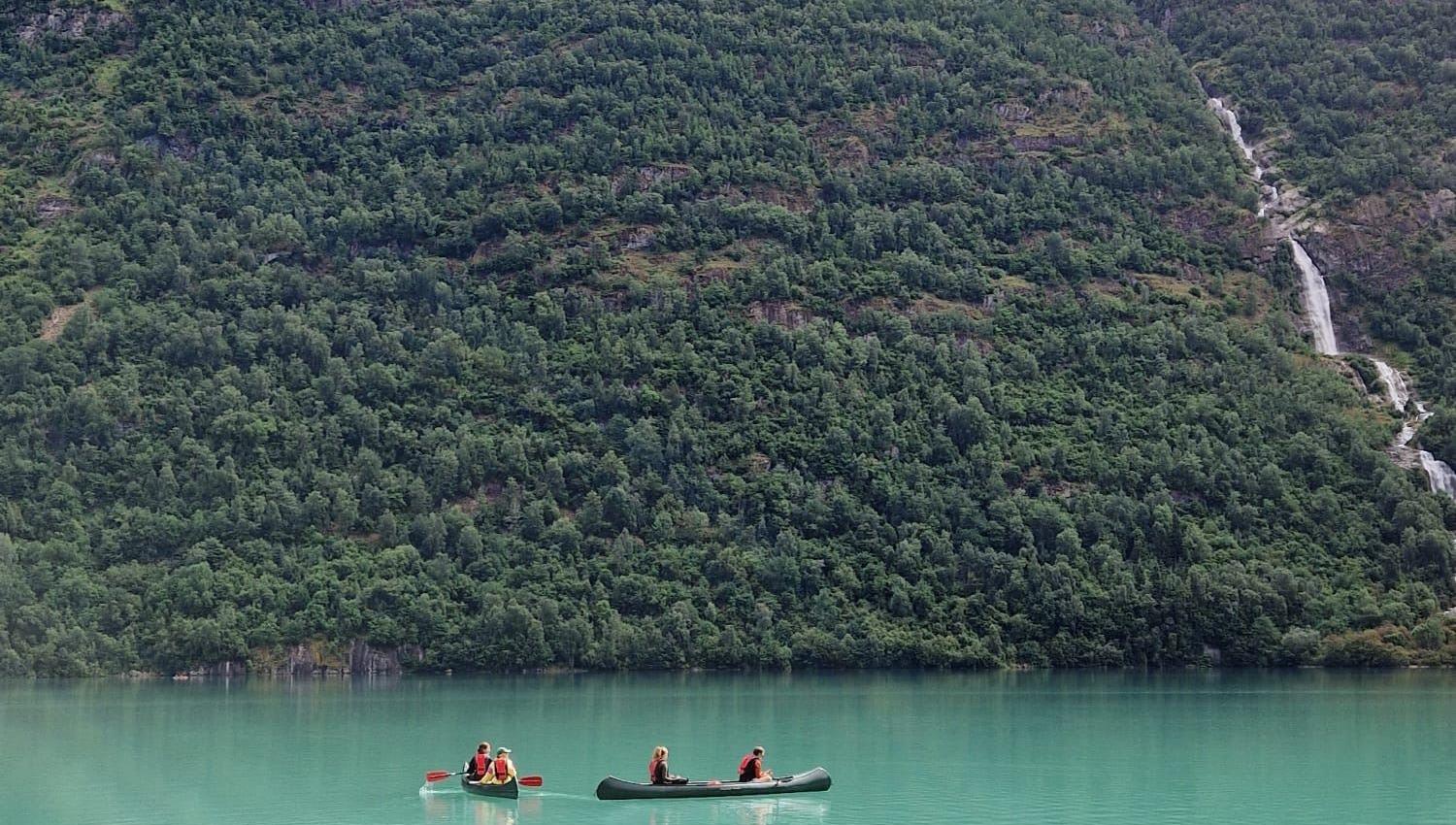 Two canoes with paddlers float on calm turquoise water below forested mountains and a distant waterfall..