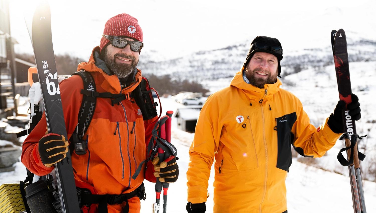 Two bundled skiers in bright jackets hold skis and poles outdoors, with snowy hills and a winter landscape behind..